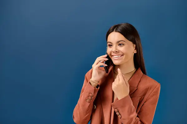 Joyeuse adolescente élégante dans un blazer à la mode parlant par téléphone et souriant joyeusement sur fond bleu — Photo de stock