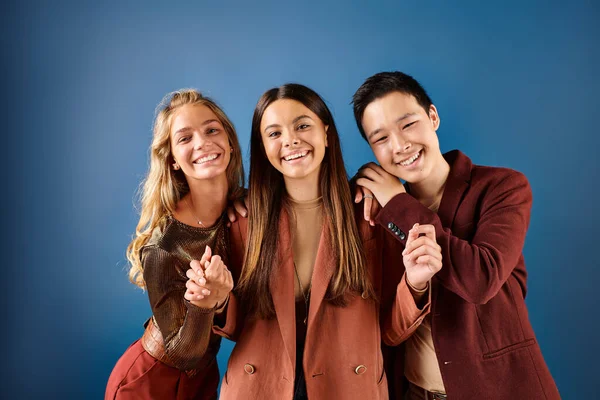 Alegres adolescentes multirraciales con ropa elegante sonriendo a la cámara en el fondo azul, día de la amistad - foto de stock