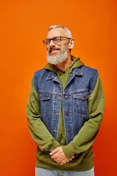 Fröhlicher flippiger Mann in grünem Kapuzenpulli und Jeansweste posiert vor orangefarbenem Hintergrund mit geschlossenen Augen — Stockfoto