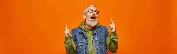 Sorpreso uomo maturo con gli occhiali e la barba in felpa con cappuccio vivido che punta su sfondo arancione, banner — Foto stock