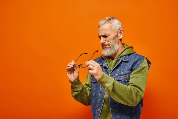 Schöner, konzentrierter, reifer Mann in lebendigem Outfit, der seine Brille in den Händen hält auf orangefarbenem Hintergrund — Stockfoto