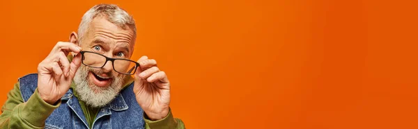 Homme mûr barbu gris joyeux en sweat à capuche vibrant et gilet en denim posant sur fond orange, bannière — Photo de stock