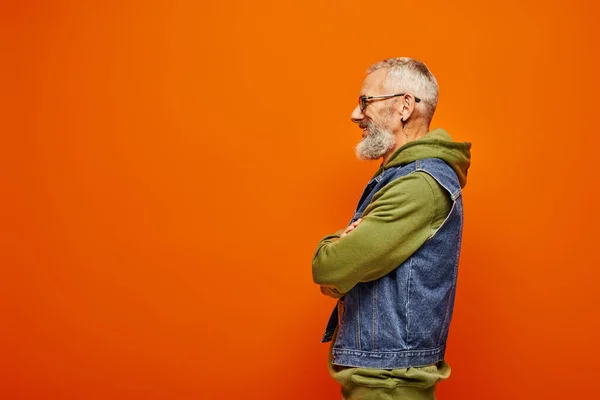 Bel homme mature joyeux en sweat à capuche vert et gilet en denim posant de profil sur fond orange — Photo de stock