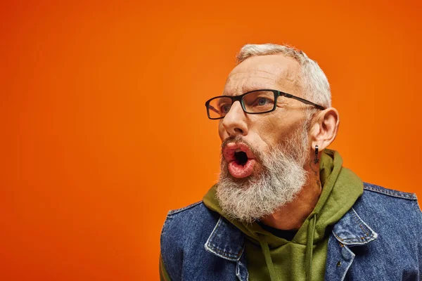 Beau mature homme en sweat à capuche vert avec des lunettes posant avec la bouche ouverte sur fond orange — Photo de stock