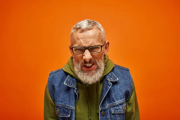 Frustrated mature emotional man in vibrant green hoodie and denim vest posing on orange background — Stock Photo