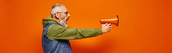 Attractive mature man in green hoodie and stylish denim vest aiming with megaphone in hands, banner — Stock Photo