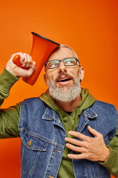 Joyeux surpris mature homme en sweat à capuche vert et gilet en denim posant avec mégaphone sur fond orange — Photo de stock