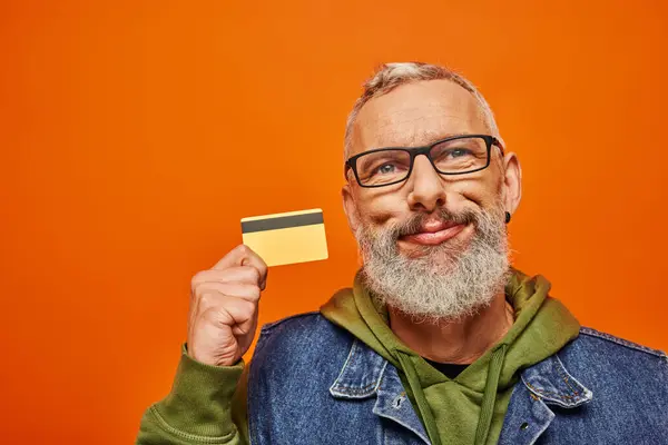 Весёлый, хорошо выглядящий зрелый мужчина в яркой одежде с очками, держащими кредитную карту у его лица — стоковое фото