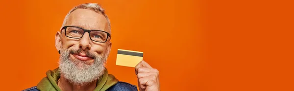 Homme mûr joliment attrayant en vêtements vifs avec des lunettes tenant la carte de crédit près de son visage, bannière — Photo de stock