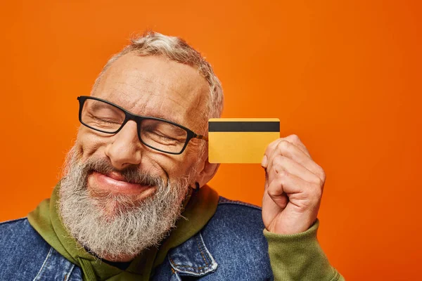Guapo modelo masculino maduro en ropa vibrante posando con los ojos cerrados y sosteniendo la tarjeta de crédito - foto de stock