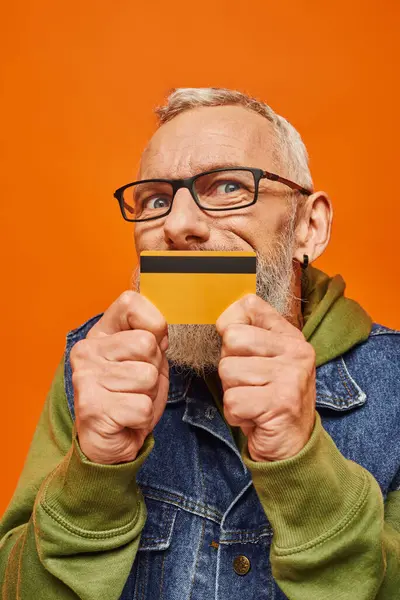 Attrayant homme mature en sweat à capuche vert et gilet en denim tenant la carte de crédit et regardant la caméra — Photo de stock