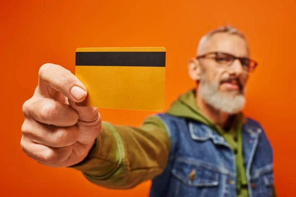 Centrarse en la tarjeta de crédito en manos de hombre maduro guapo borrosa en sudadera con capucha vibrante mirando a la cámara - foto de stock