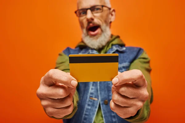 Se concentrer sur la carte de crédit dans les mains de l'homme barbu flou mature en tenue vibrante sur fond orange — Photo de stock