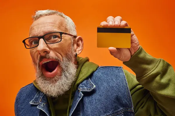 Joyeux beau modèle masculin mature en tenue vibrante tenant carte de crédit sur fond orange — Photo de stock