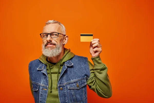 Alegre atractivo hombre maduro en chaleco de mezclilla con gafas mirando a la tarjeta de crédito en el fondo naranja - foto de stock
