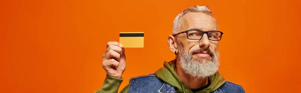 Alegre guapo maduro hombre en denim chaleco y verde sudadera con capucha mirando tarjeta de crédito en la mano, bandera - foto de stock