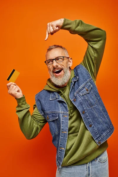 Веселый привлекательный зрелый мужчина модель в стильном джинсовом жилете указывая на кредитную карту в его руках — стоковое фото
