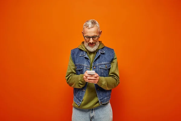 Bello funky uomo maturo in verde felpa con cappuccio alla moda guardando smartphone su sfondo arancione — Foto stock