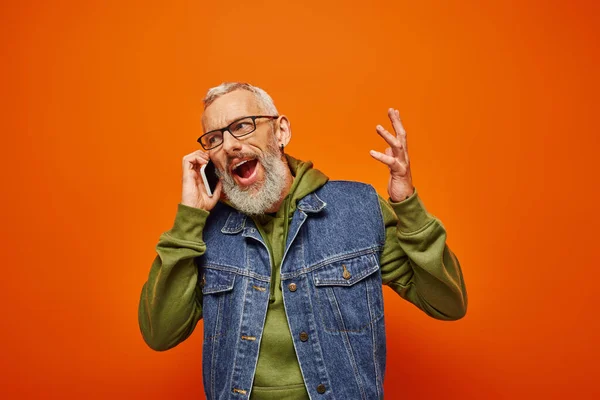 Красивый радостный зрелый мужчина в ярко-зеленой толстовке и джинсовой жилетке в очках разговаривает по телефону — стоковое фото