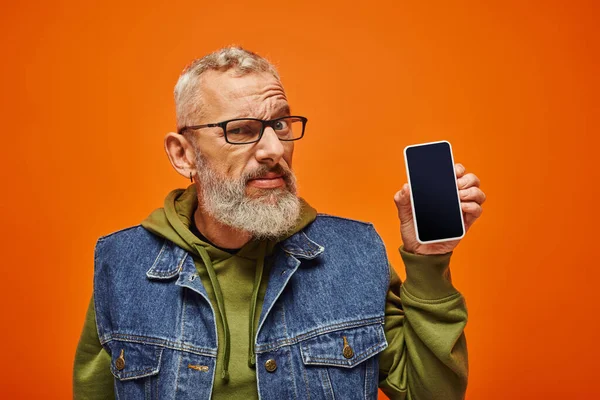 Verwirrter attraktiver älterer Mann in leuchtend grünem Kapuzenpulli, Telefon in der Hand und in die Kamera blickend — Stockfoto