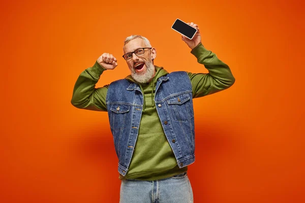 Joyeux attrayant homme mature en sweat à capuche vert et gilet en denim tenant le téléphone et regardant la caméra — Photo de stock