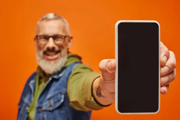 Se centran en el teléfono inteligente en manos del hombre maduro alegre borrosa en ropa vibrante sobre fondo naranja - foto de stock