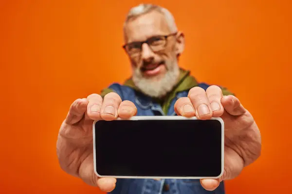 Se centran en el teléfono inteligente en manos de un hombre alegre y maduro borroso con un atuendo vibrante sobre un fondo naranja - foto de stock