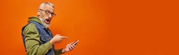Homme mature attrayant choqué en sweat à capuche vert pointant vers le smartphone et regardant la caméra, bannière — Photo de stock