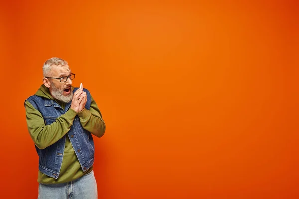 Attraktives reifes männliches Modell in grünem Kapuzenpulli und Jeansweste, das per Smartphone auf orangefarbenem Hintergrund spricht — Stockfoto