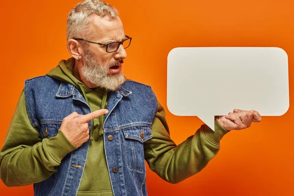 Verwirrter gutaussehender älterer Mann in Jeansweste blickt auf Sprechblase in der Hand vor orangefarbenem Hintergrund — Stockfoto
