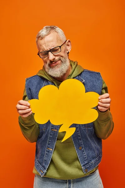 Verwirrter gereifter Mann in Jeansweste blickt auf Gedankenblase in seinen Händen vor orangefarbenem Hintergrund — Stockfoto