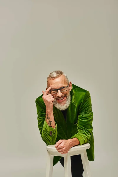 Homem maduro barbudo alegre em trajes vívidos com tatuagens posando com cadeira alta e sorrindo para a câmera — Fotografia de Stock