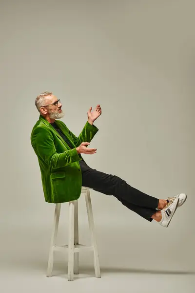 Joyous mature man in green blazer with glasses sitting on tall chair in profile on beige backdrop — Stock Photo