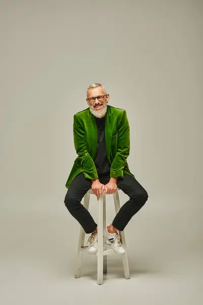 Alegre modelo masculino funky con barba y gafas sentado en una silla alta y sonriendo a la cámara - foto de stock