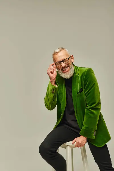 Bien parecido hombre funky en chaqueta verde sentado en silla alta y sonriendo sinceramente con los ojos cerrados - foto de stock