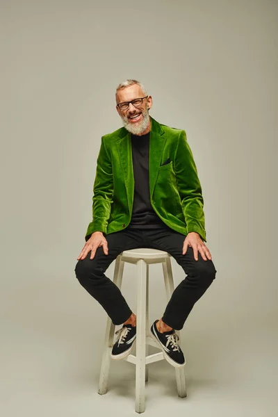 Homem maduro atraente barbudo alegre em blazer verde sentado em cadeira alta e sorrindo para a câmera — Fotografia de Stock