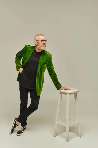 Homem maduro de boa aparência com barba em traje vibrante posando ao lado de cadeira alta e olhando para longe — Fotografia de Stock