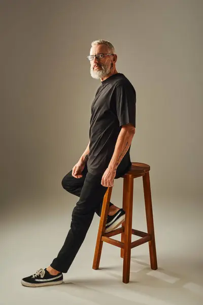 Schöner älterer Mann in schwarzem T-Shirt sitzt auf einem hohen Stuhl und blickt in die Kamera auf beigem Hintergrund — Stockfoto