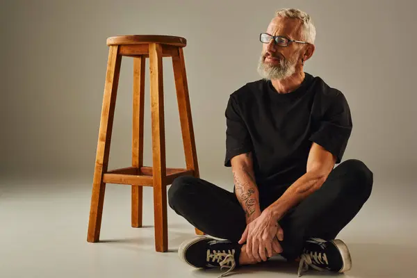 Attraktive ältere fröhliche Mann in schwarzem T-Shirt mit Tätowierungen sitzt auf dem Boden mit überkreuzten Beinen — Stockfoto