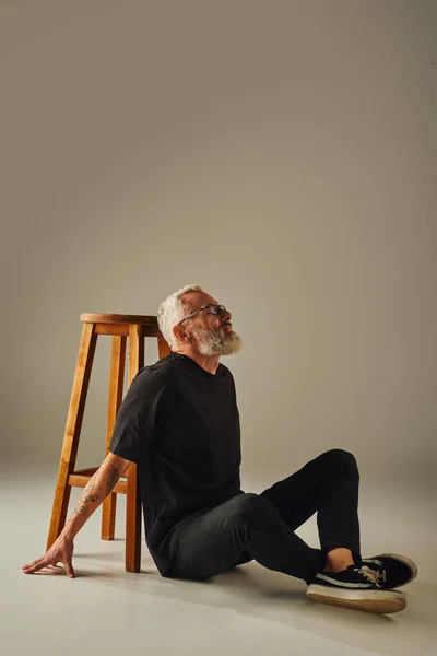 Modelo masculino maduro de buen aspecto en camiseta negra con gafas sentadas en el suelo junto a una silla alta - foto de stock