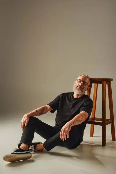 Attraktiver, reifer Mann in schwarzem T-Shirt mit Bart sitzt auf dem Boden in der Nähe eines hohen Stuhls mit geschlossenen Augen — Stockfoto