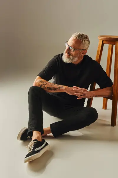 Atractivo hombre maduro en camiseta negra con gafas y tatuajes sentado en el suelo junto a una silla alta - foto de stock
