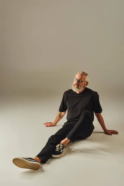 Fröhlicher reifer Mann in schwarzem T-Shirt mit Brille auf dem Boden sitzend und vor beigem Hintergrund wegschauend — Stockfoto