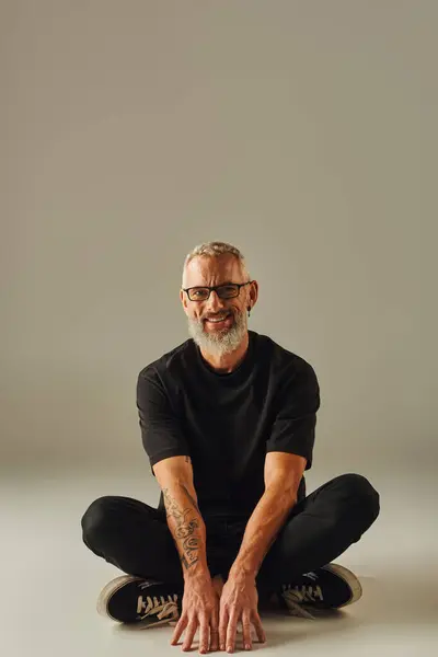 Fröhlich gut aussehender älterer Mann in schwarzem T-Shirt sitzt mit überkreuzten Beinen auf dem Boden und lächelt in die Kamera — Stockfoto