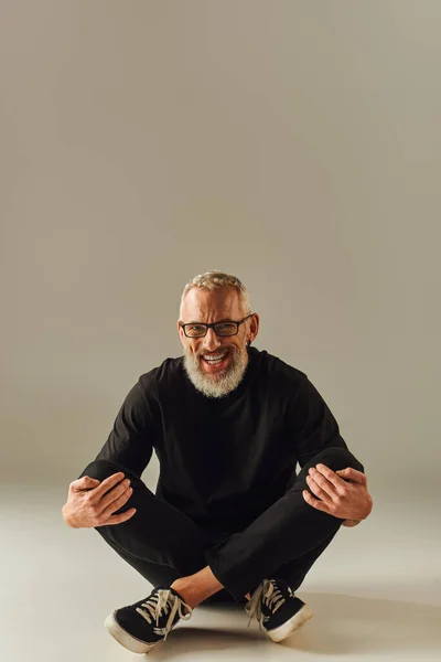 Beau gai mature homme en t-shirt noir avec des lunettes assis sur le sol avec des jambes croisées — Photo de stock