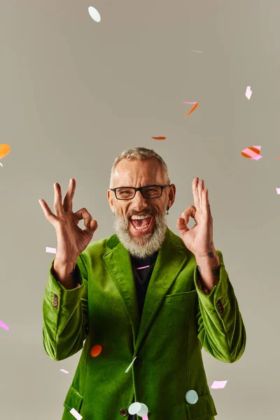 Beau modèle masculin joliment mature en veston vert montrant un signe acceptable à la caméra sous la pluie confettis — Photo de stock