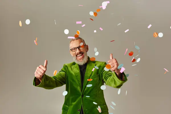 Alegre modelo masculino maduro em blazer verde mostrando polegares para cima sob chuva confete no fundo bege — Fotografia de Stock