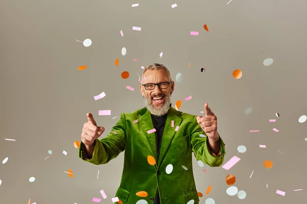Joyeux beau mature homme en vert blazer pointant vers la caméra sous la pluie confettis sur fond beige — Photo de stock
