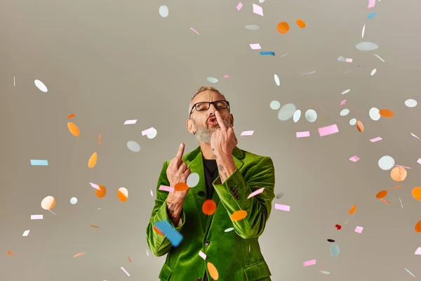 Cheeky mature man in green vibrant blazer showing middle fingers at camera under confetti rain — Stock Photo