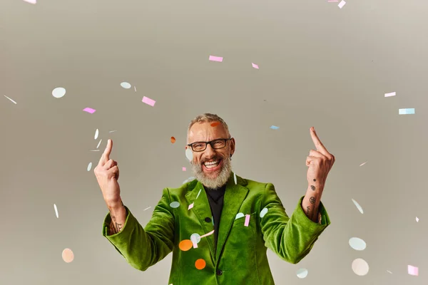 Alegre hombre maduro en verde chaqueta vibrante con gafas mostrando los dedos medios y sonriendo a la cámara - foto de stock